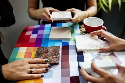 Three people sitting around a colorful table, painting tiles.